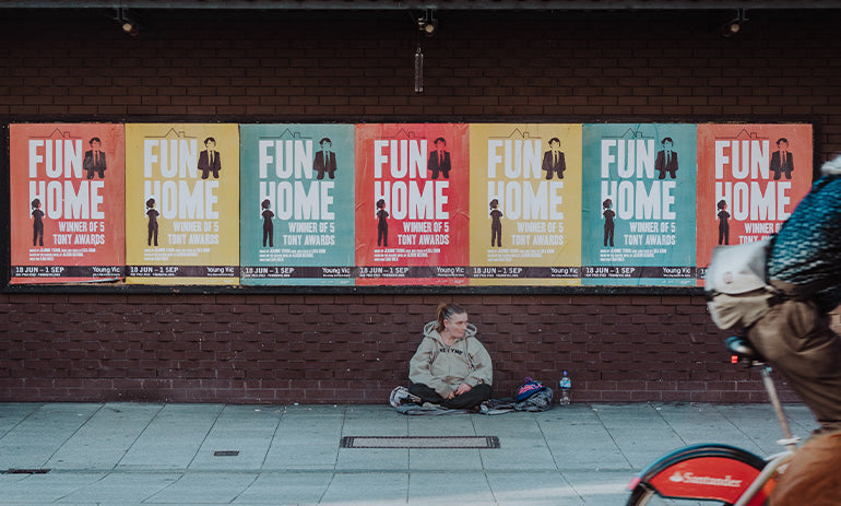 A woman sits on a street path beneath colourful posters as a bike rides past. It is apparent that she is experiencing homelessness.