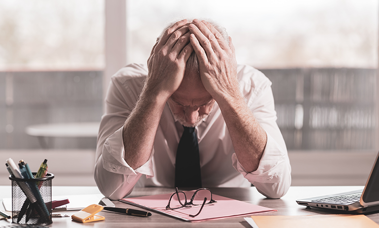 Older man slumped in office chair with his head in his hands, wearing a suit.
