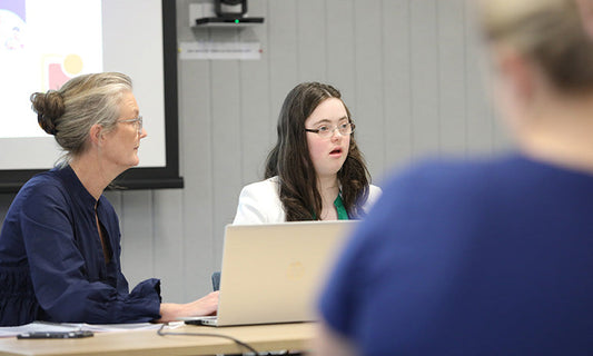 A dark-haired lady sits at a table, giving a presentation. She has Down syndrome. An older lady with grey hair sits next to her.|A woman holds a stethoscope cord in the shape of a heart. We can see her torso and arms. She is wearing blue scrubs.