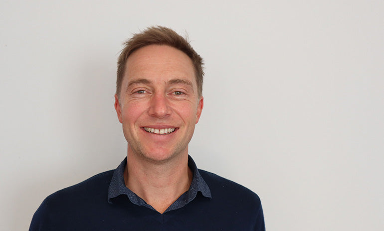 A man in a dark coloured jumper with short brown hair is smiling at the camera.