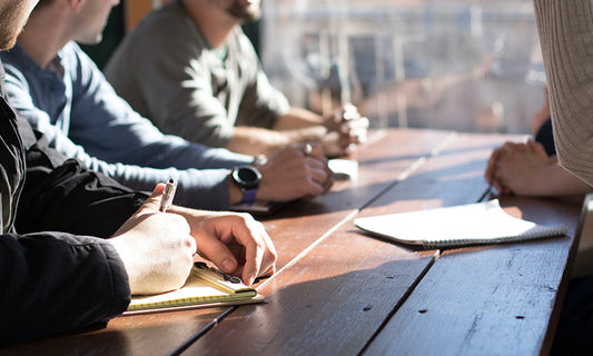 A group of people sit around a table. They are all dressed casually, but we can't see their faces, only their hands and arms. There is also paper on the table, and it is sunny through the window.