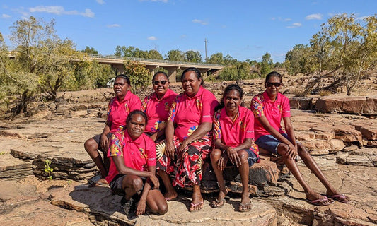 A group of Aboriginal women in pink uniforms sit outside on Country. They are Indi Kindi educators.|Three Aboriginal kids sit on a blanket on country. They look like they are listening to someone.
