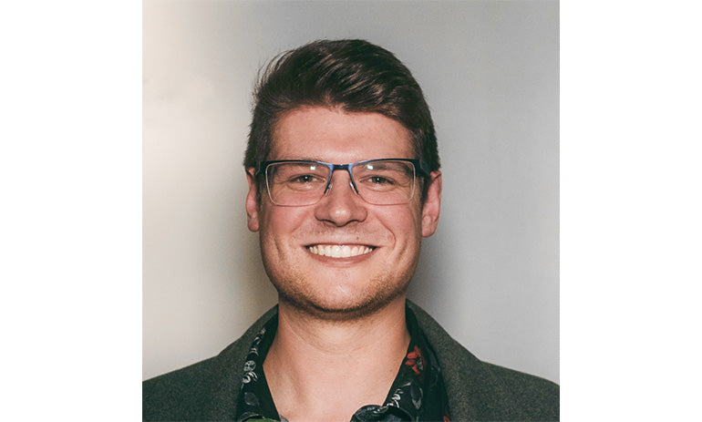 Joshua Morley, a white man, smiles at the camera. He has brown hair and wears glasses. We can see him from the shoulders up. He is wearing dark coloured clothes.