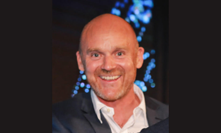 Korey Allchin smiles at the camera. He is wearing a suit jacket and white shirt and is bald. We can see him from the shoulders up.