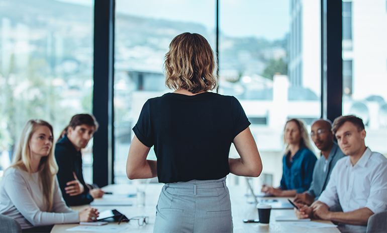 Back of young woman addressing a room of colleagues.