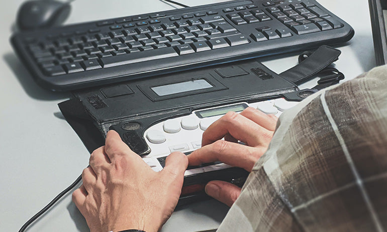 A keyboard in the background as an accessibility keyboard attached to it. A person is using the accessible keyboard. We are looking over their shoulder and can only see their hands.