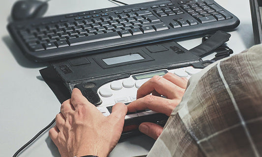 A keyboard in the background as an accessibility keyboard attached to it. A person is using the accessible keyboard. We are looking over their shoulder and can only see their hands.