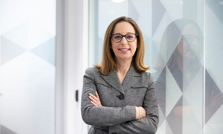 A lady smiles at the camera, her arms folded. She is wearing glasses and has light brown hair down to her shoulders.