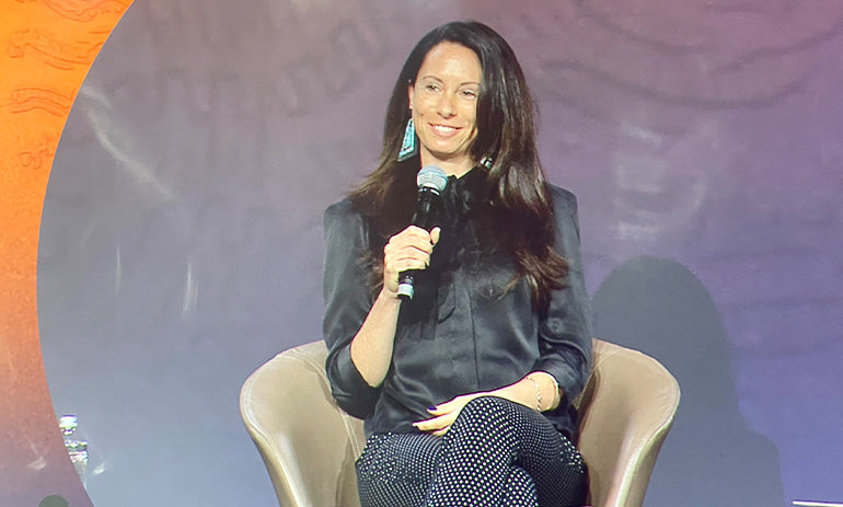 Tara Anderson, a white lady with dark hair, sits on a stage holding a microphone. She is wearing black and is smiling.