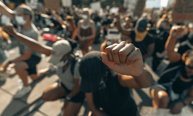 A group of people protesting racism. In the foreground a young person has their fist raised.||Wellcome|Sonia Safat, an Indian woman of colour, smiles at the camera. She has brown hair and is wearing a blue denim shirt.