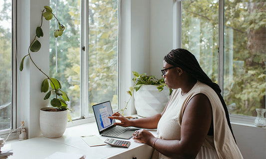 A Black woman sits at a desk with her laptop. There are plants on the desk and the room is white and peaceful looking.