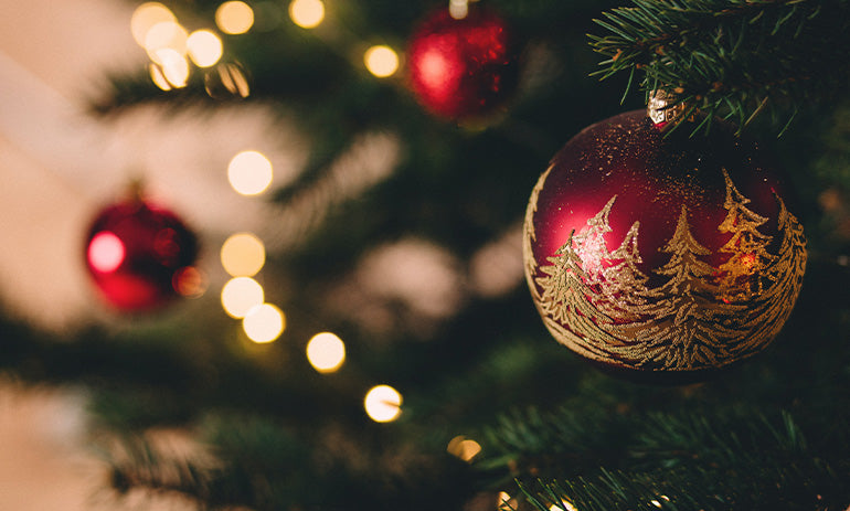A red Christmas bauble on a Christmas tree. It has gold Christmas trees painted on it, and we can see other baubles in the background.