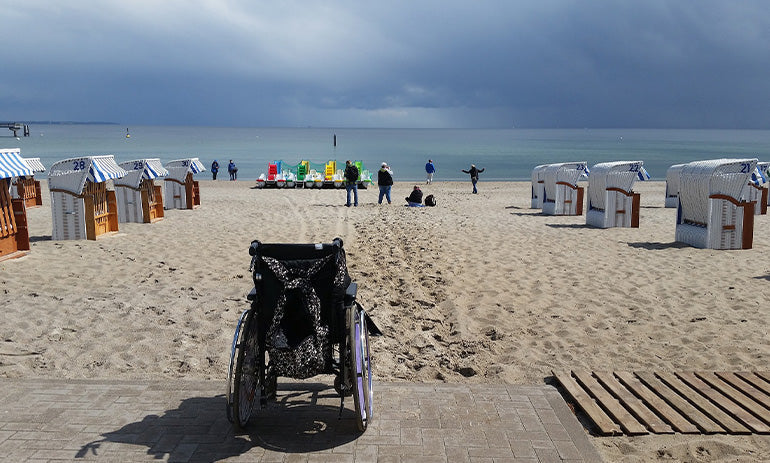 A beach scene, with huts along the sand. In the foregound is a wheelchair, facing out towards the water. There are people on the sand in the distance.