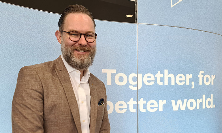 Ben Cox, a white man with a greying beard and moustache, brown hair and black glasses, smiles at the camera. He is at a conference, standing in front of a blue board that says 'Together, for a better world'. He is wearing a brown blazer and white shirt.|
