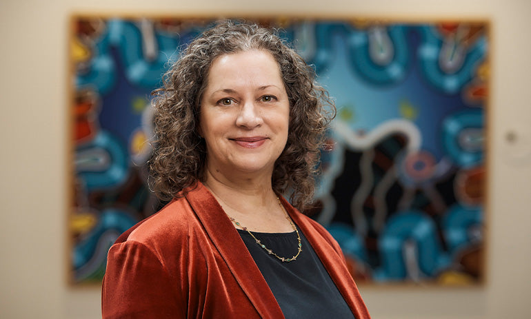 Kelly Bruce smiles at the camera. She is standing in front of a colourful painting and wearing an orange top. She has brown curly hair.