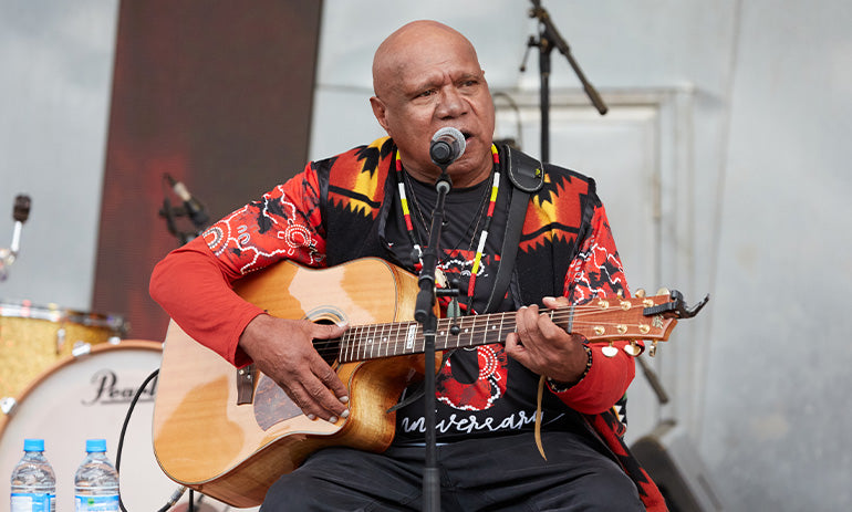 Archie Roach, an Aboriginal man, sits behind a microphone playing an acoustic guitar. He is wearing a shirt i the colours of the Aboriginal flag: red, yellow and black.