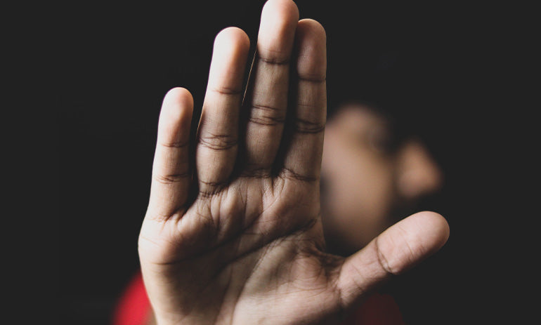 A person on a dark background holds up their hand in a stop sign. They are blurred out in the background.