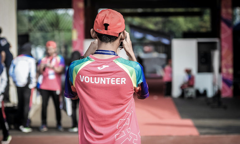 We see a person from behind. They are wearing a red and blue shirt with the word volunteer on the back, and a red cap. They are standing in front of a building and some people.
