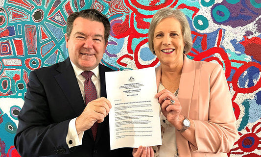 Senators Dean Smith and Catryna Bilyk smile at the camera. They are holding a piece of paper between them with words we can't read.