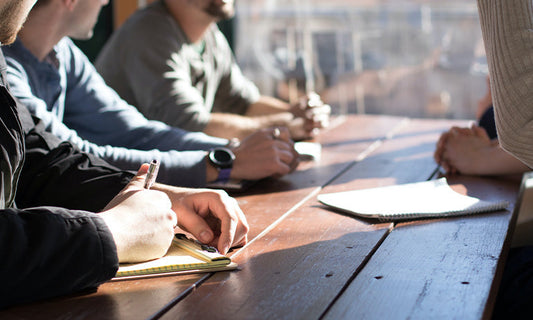 A group of people sitting around a table talking and writing notes. We can only see their torsos and hands.|Disability employment