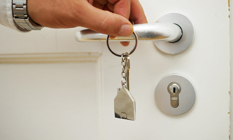 We see a door handle and a lock. There is a key ring hanging off the handle with a silver house on it. A person is touching the handle and we see their hand.