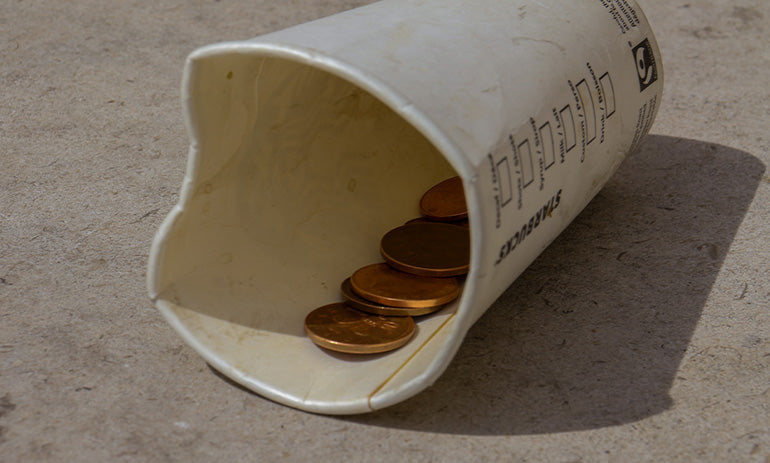 A white cup, perhaps an old coffee cup, lying on a pale surface. There are some gold coins inside. It looks like it may have been used by a person experiencing homelessness to collect money.
