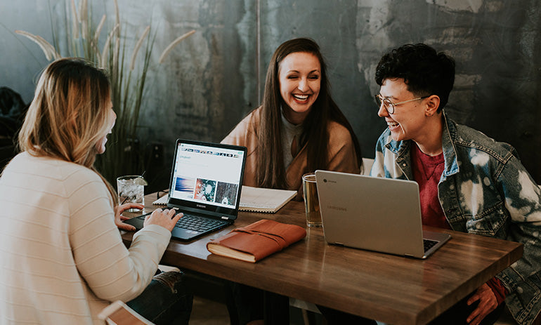 A group of three work colleagues sit around a table with their laptops. They are all laughing and dressed casually.