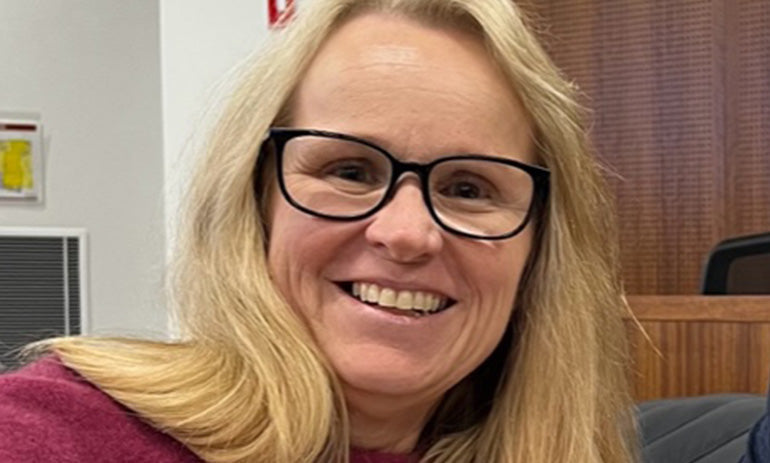 A lady smiles at the camera. She has long blonde hair and black rimmed glasses.