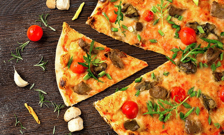 A delicious looking pizza on a stone background. The pizza has tomatoes, rocket, cheese andother vegetables and has a slice taken out of it and moved slightly to be next to it. On the board next to the pizza are rosemary, tomatoes and mushrooms.
