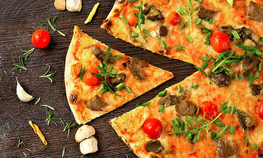 A delicious looking pizza on a stone background. The pizza has tomatoes, rocket, cheese andother vegetables and has a slice taken out of it and moved slightly to be next to it. On the board next to the pizza are rosemary, tomatoes and mushrooms.