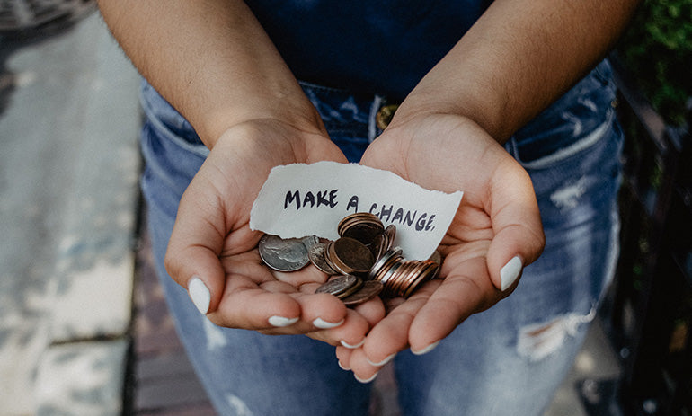 We are looking down at a person from above. We can see their blue jeans and outstretched hands holding coins and a little sign that says 'make a change'.
