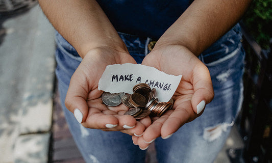 We are looking down at a person from above. We can see their blue jeans and outstretched hands holding coins and a little sign that says 'make a change'.