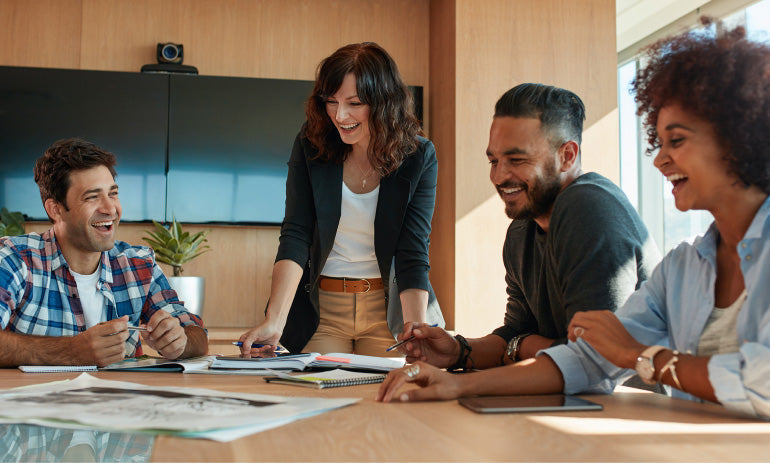 A group of people sit around a table spread with documents. They are all laughing.