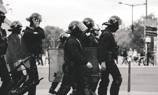 A black and white image of police in riot gear walking in a group down the street.