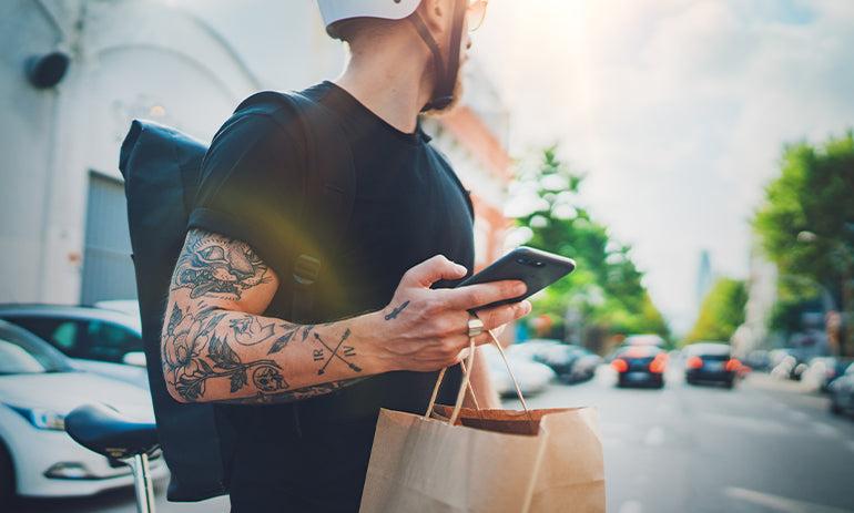 A man stands in front of the camera, turned away from it. He has tattoors on his arm and is holding a brown paper bag and his phone. He is a gig worker, delivering food.|