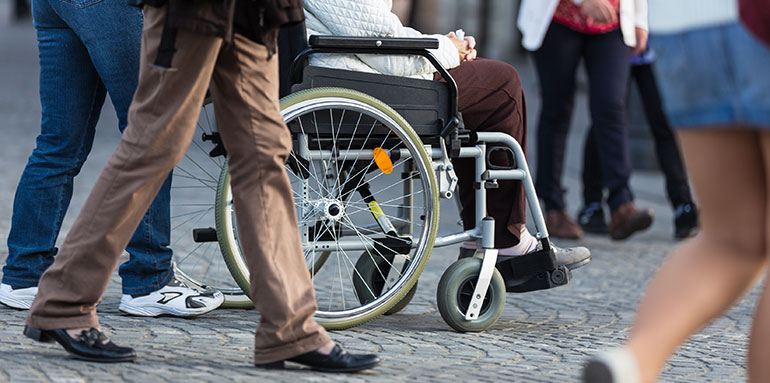 A picture showing the lower half of people walking around, and a person pushing another in an wheelchair in the centre of the photo. No faces can be seen only legs and wheels.
