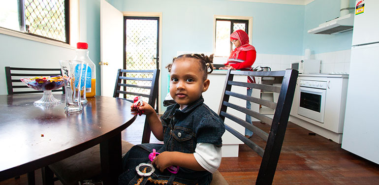 Young girl sitting at kitchen table with her mum in the background.