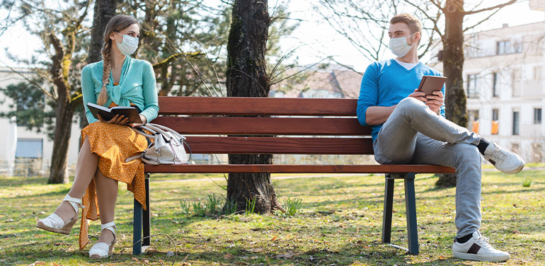 man and a woman wearing facemasks sitting at either end of a park bench with a space between them