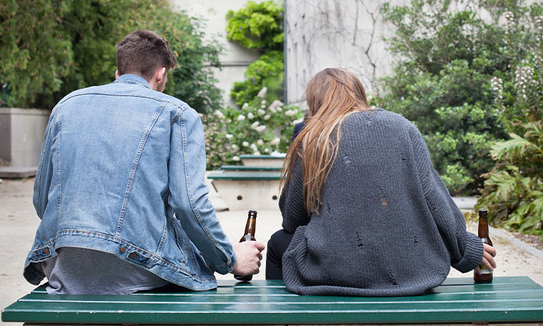 young man and woman with backs to the camera sitting on a bench drinking beer