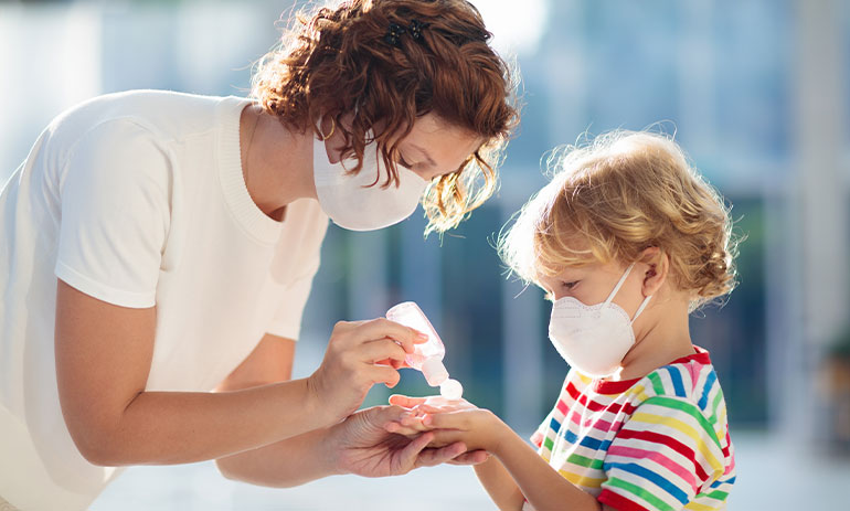 woman putting hand sanitiser on child's hands, both wearing masks.