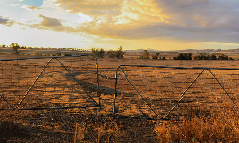 Panoramic views of dry, drought stricken farm land through old steel locked farm gates on a hot afternoon in rural Australia.