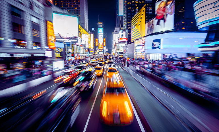 Blurred image of cars driving through Times Square with billboards visible in the background