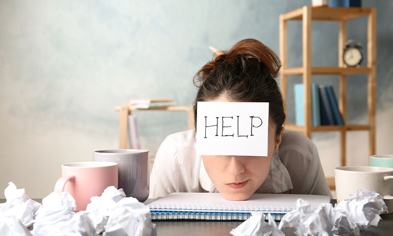 woman with chin on her desk with the word help on a piece of paper stuck to her forehead