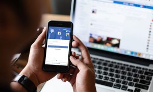 Person holding phone with facebook page on the screen, laptop in the backgroup also shows Facebook profile page