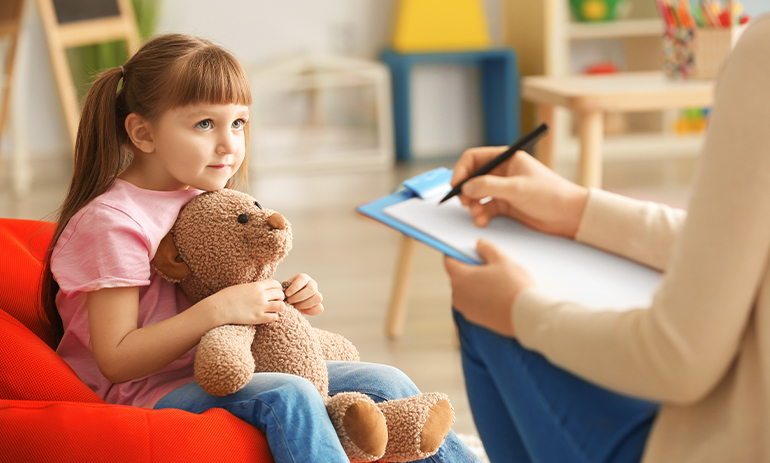 young girl holding a teddy bear looking at a counsellor holding a clipboard