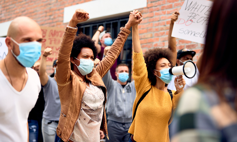 Multi-ethnic crowd of people wearing protective face mask while protesting on anti-racism demonstrations. Focus is on two black women holding hands.