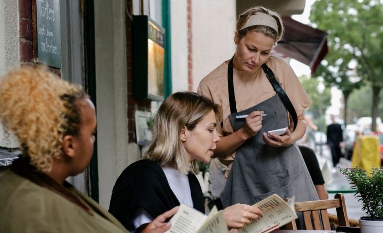 two women sitting at an outside table in a restaurant ordering from a waitress