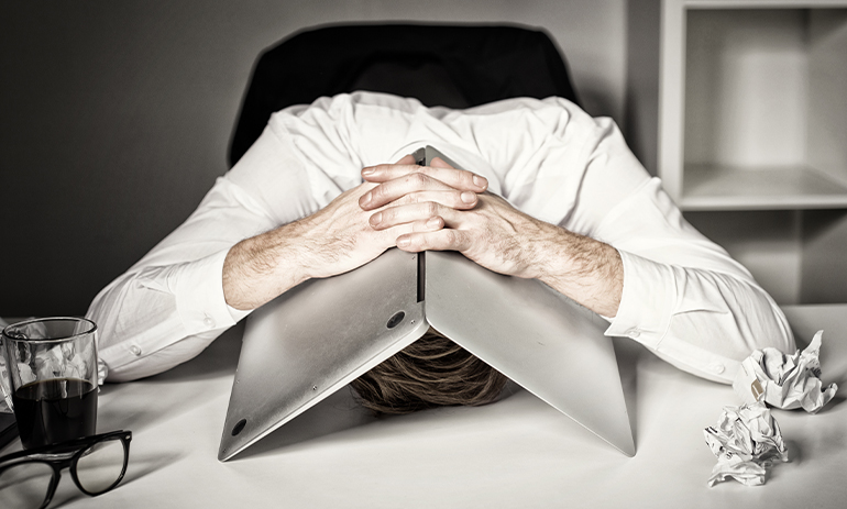 man with head on his desk with his laptop covering his head