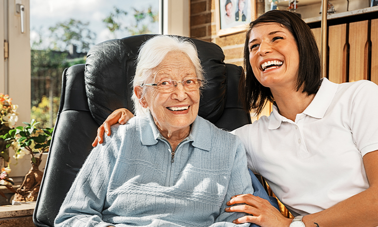 Smiling older woman sat in a chair next to younger female carer