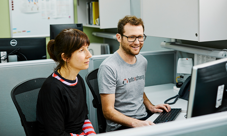 a man and a woman sitting next to each other looking at a computer. The man is wearing an Infoxchange t-shirt